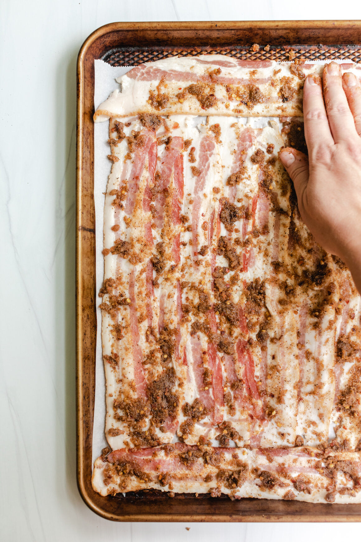 Hand spreading sugar and spice mixture over bacon on a baking sheet to make Million Dollar Bacon