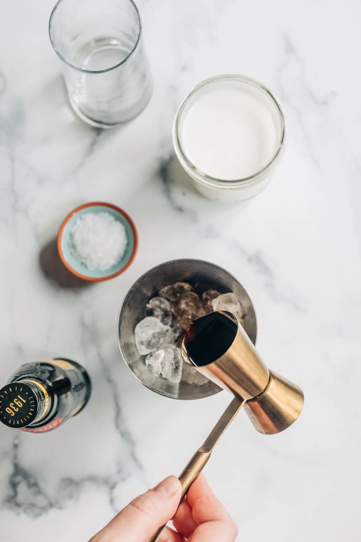Female hand pouring Kahlua into a cocktail shaker over ice using a brass jigger